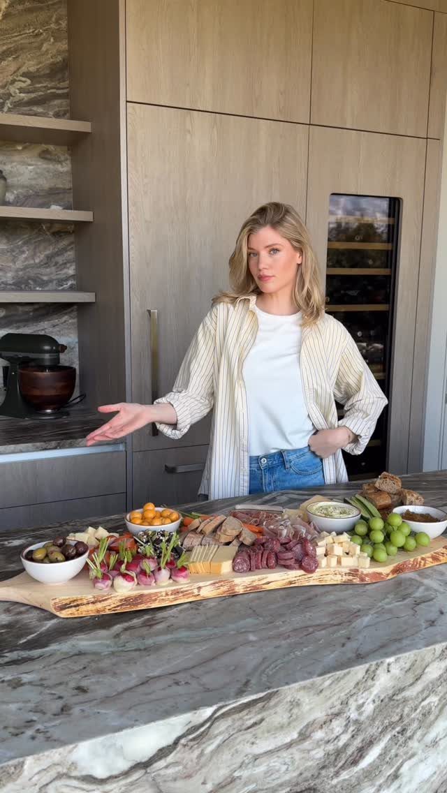 Woman presenting a large wooden charcuterie board with meats and cheeses on marble.