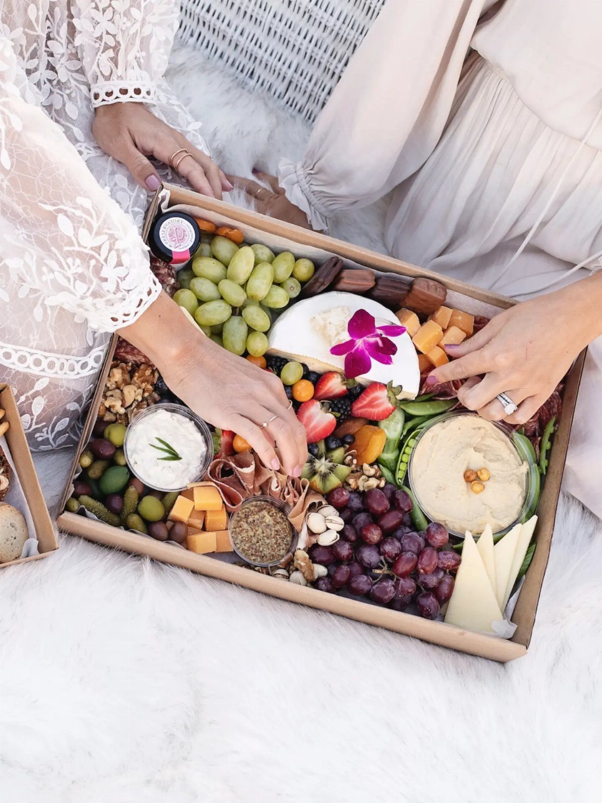 Charcuterie box with grapes, cheese, and hummus being shared by two women.