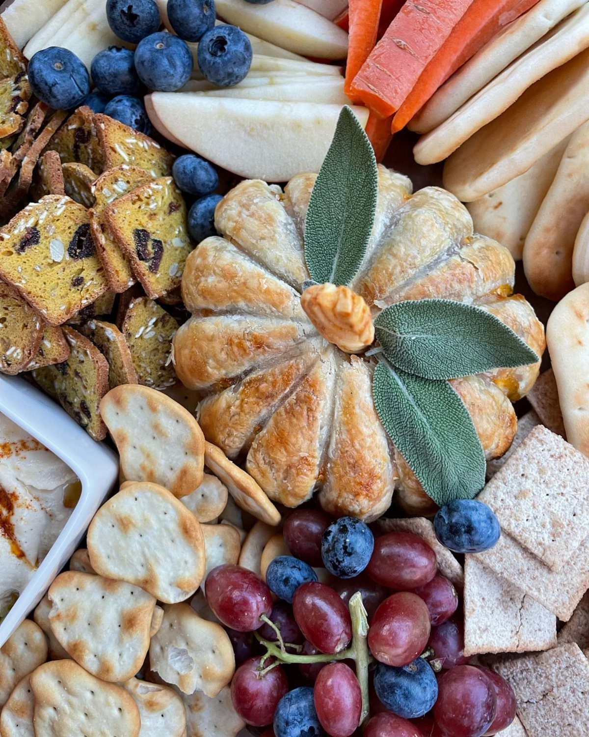 Pumpkin-shaped pastry surrounded by grapes, blueberries, crackers, and sliced fruit on a board.