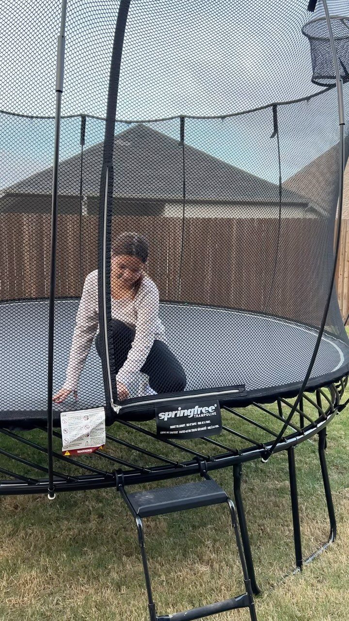 Young person sitting on a large black trampoline with a safety net outdoors.