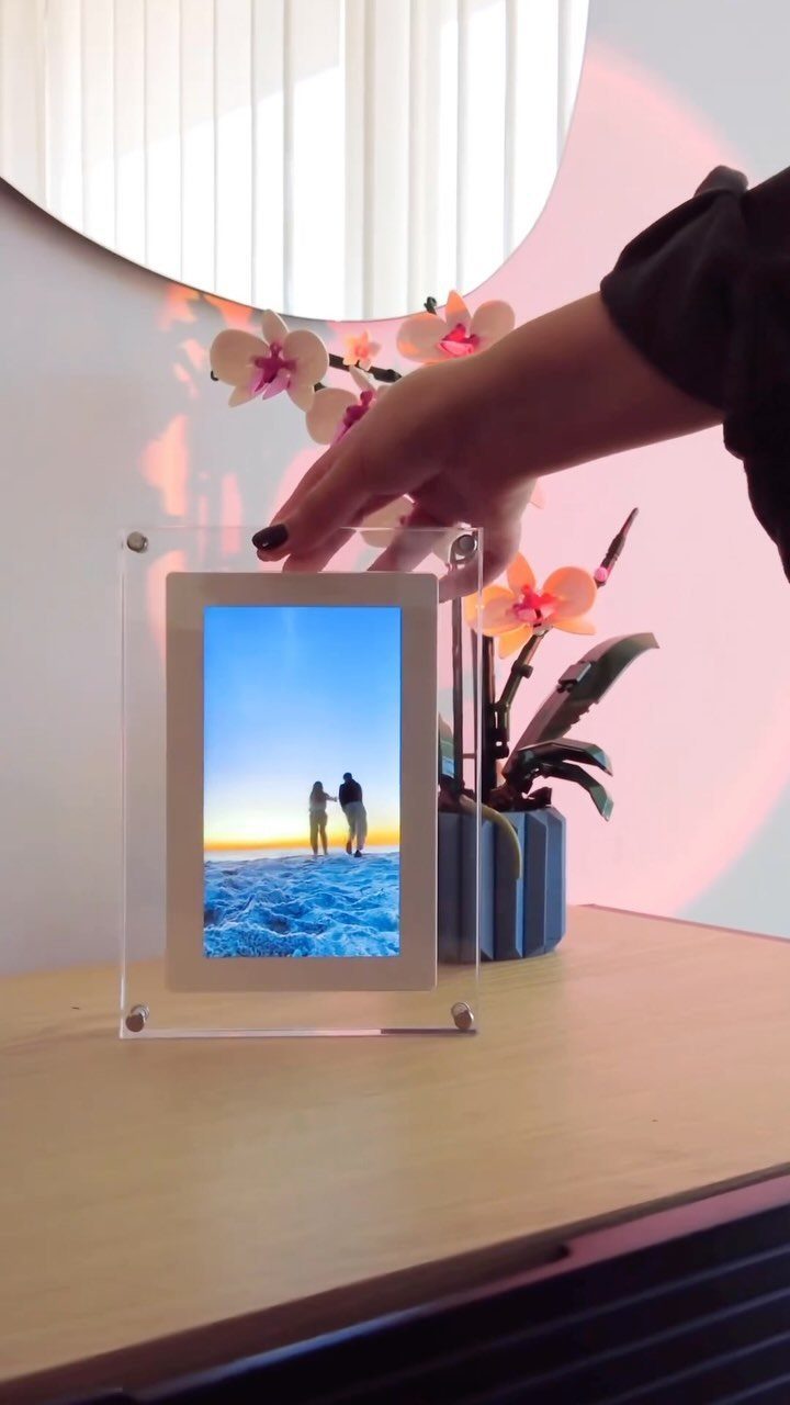 Digital picture frame displaying a beach photo on a wooden table with orchids.