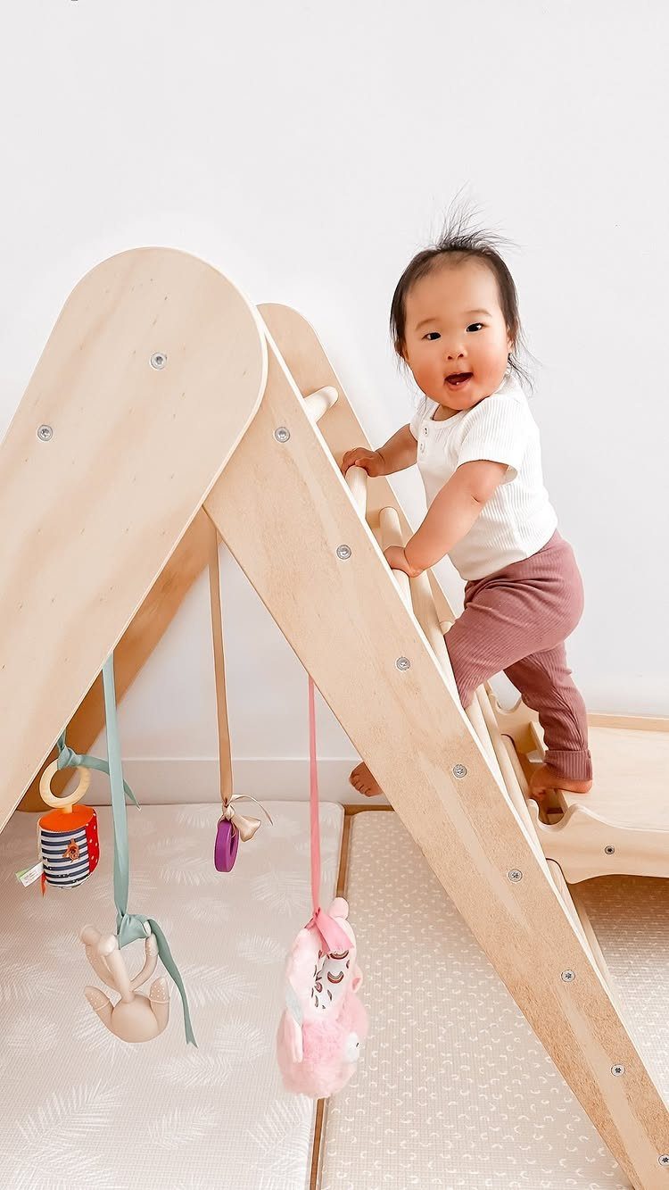 Toddler climbing a wooden Pikler triangle indoor playground with hanging toys.