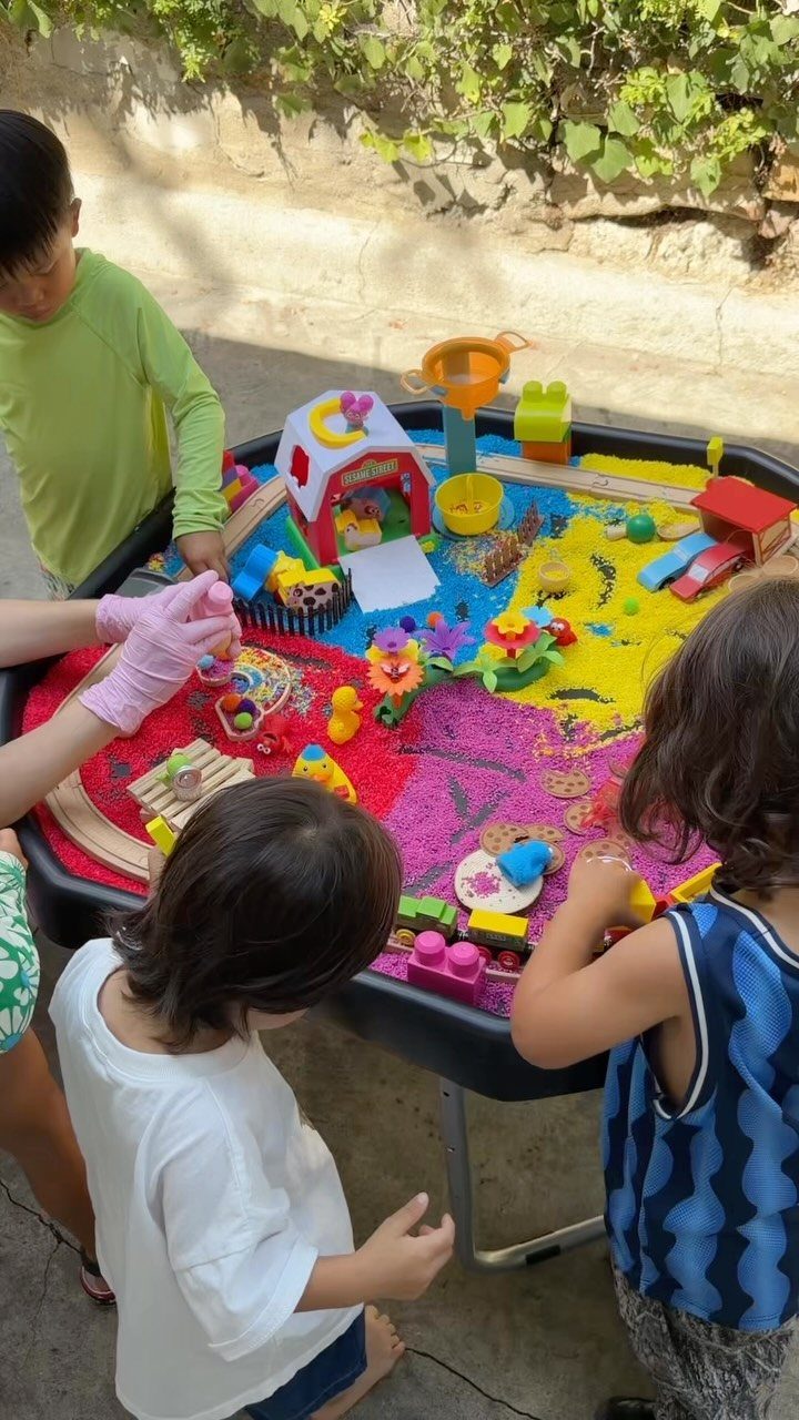 Children playing with colorful materials and toys in a round sensory table outdoors.