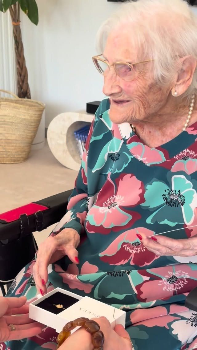 Elderly woman in floral dress receiving a gold ring in a jewelry box.