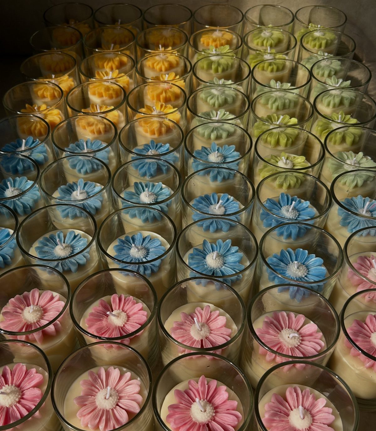 Colorful wax flower candles in small glass jars arranged in neat rows.