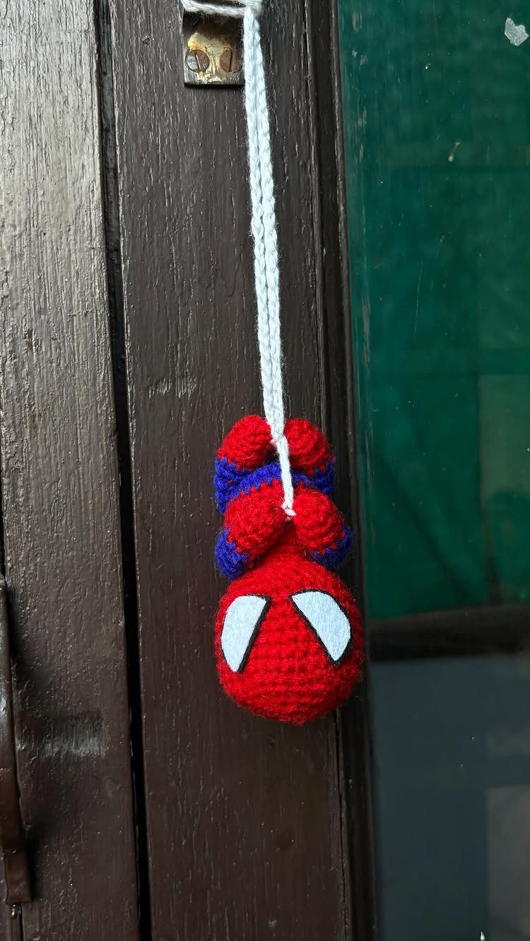Crocheted Spider-Man figure hanging by a white string from a wooden door frame.