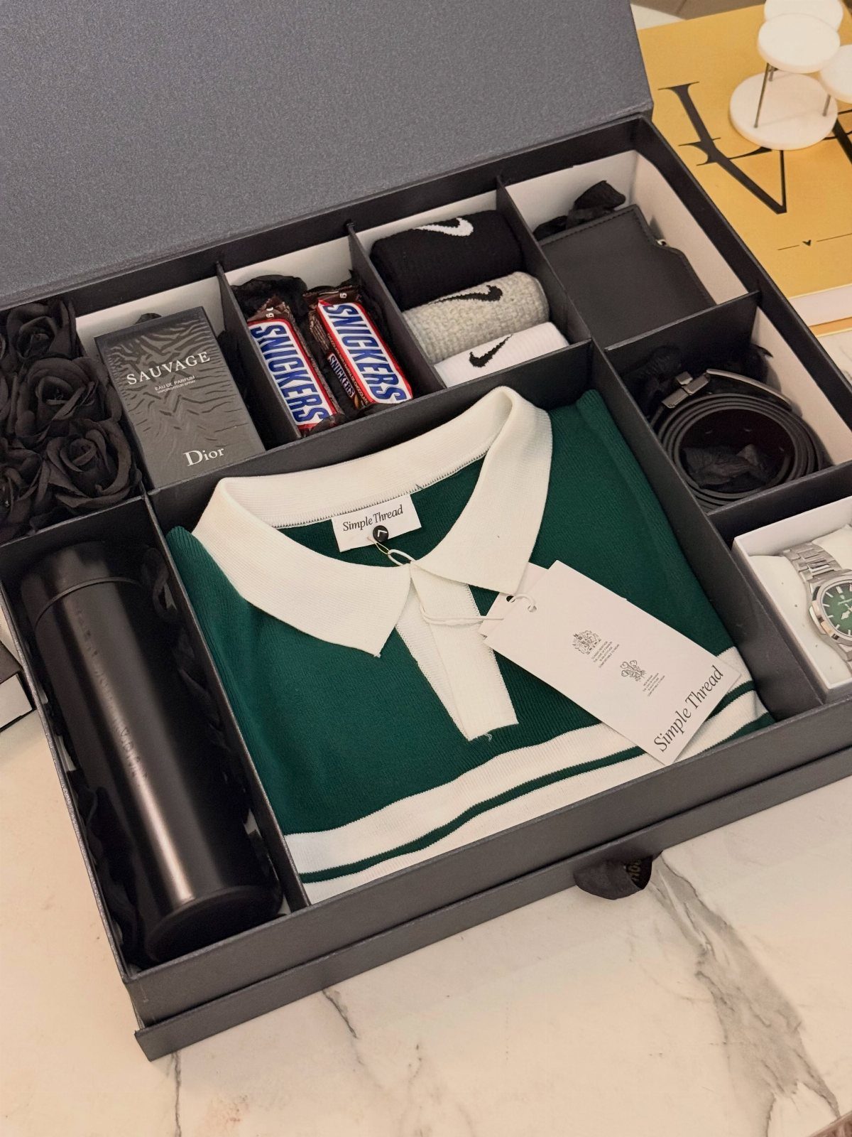 Curated gift box with green polo shirt, Dior perfume, and snacks on marble.