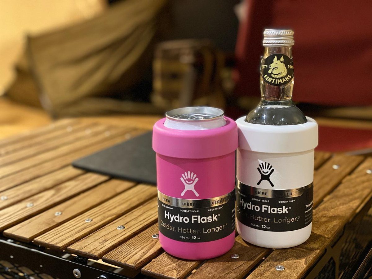 Two Hydro Flask cooler cups in pink and white holding drinks on wood.