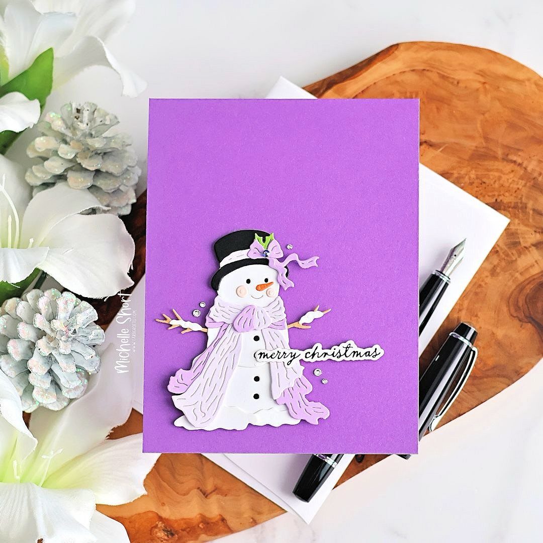 Purple Christmas card with a 3D snowman cutout, white flowers, and black pens.