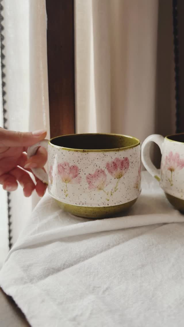 Hand holding a white ceramic mug painted with pink flowers and green base.