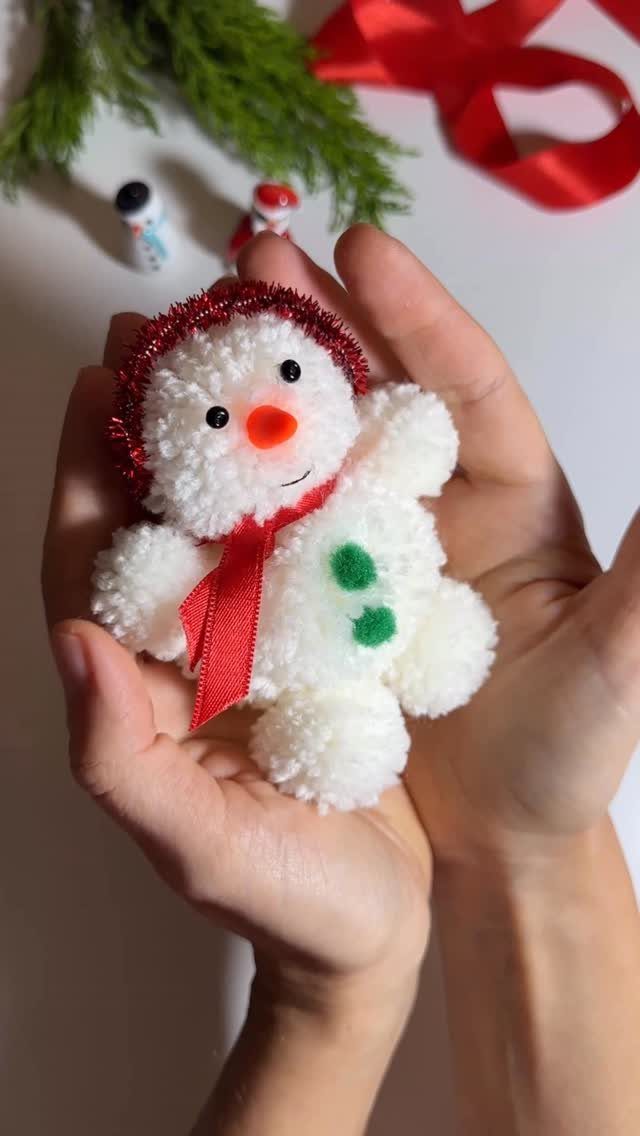 Small white pom-pom snowman with red scarf and tinsel hat held in hands