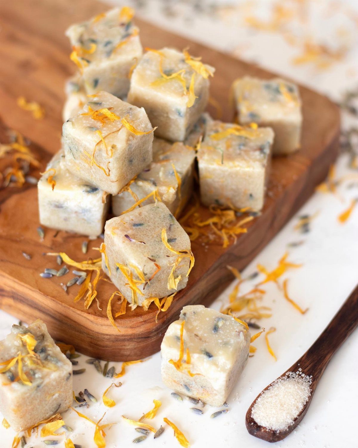 Sugar scrub cubes with dried flower petals on a wooden board and spoon.