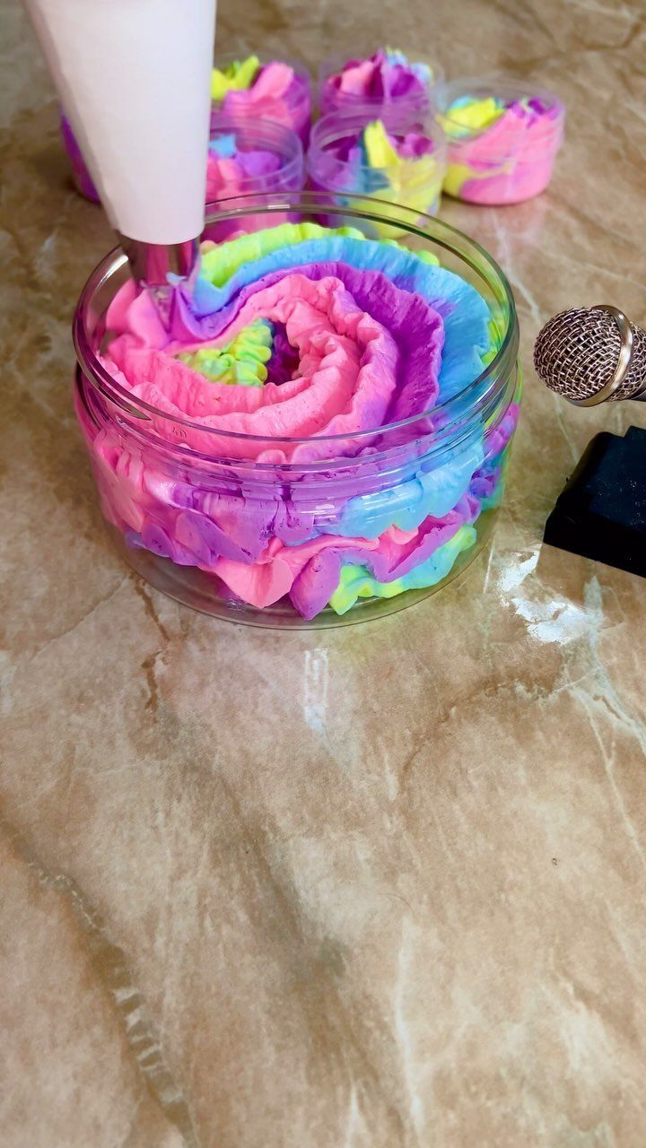 Colorful whipped body butter being piped into a clear plastic jar on marble.