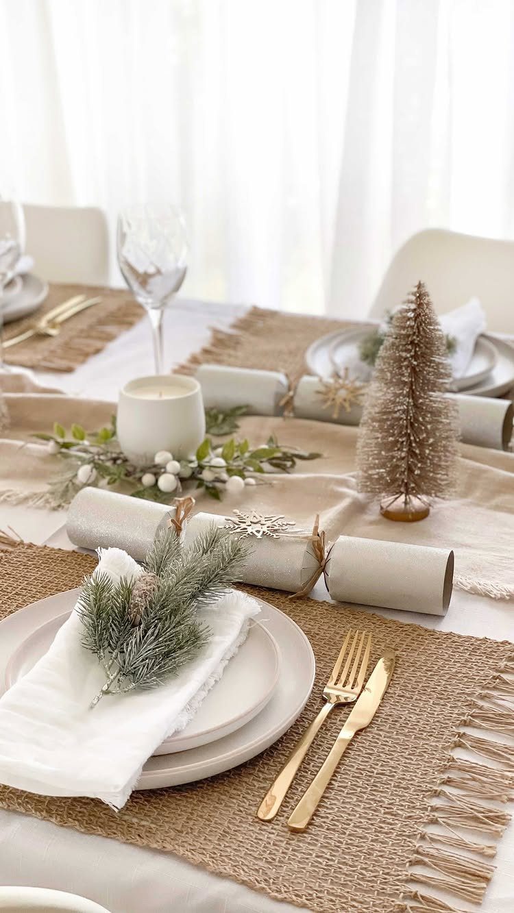 Festive table setting with white plates, gold cutlery, white candles, and evergreen greenery.
