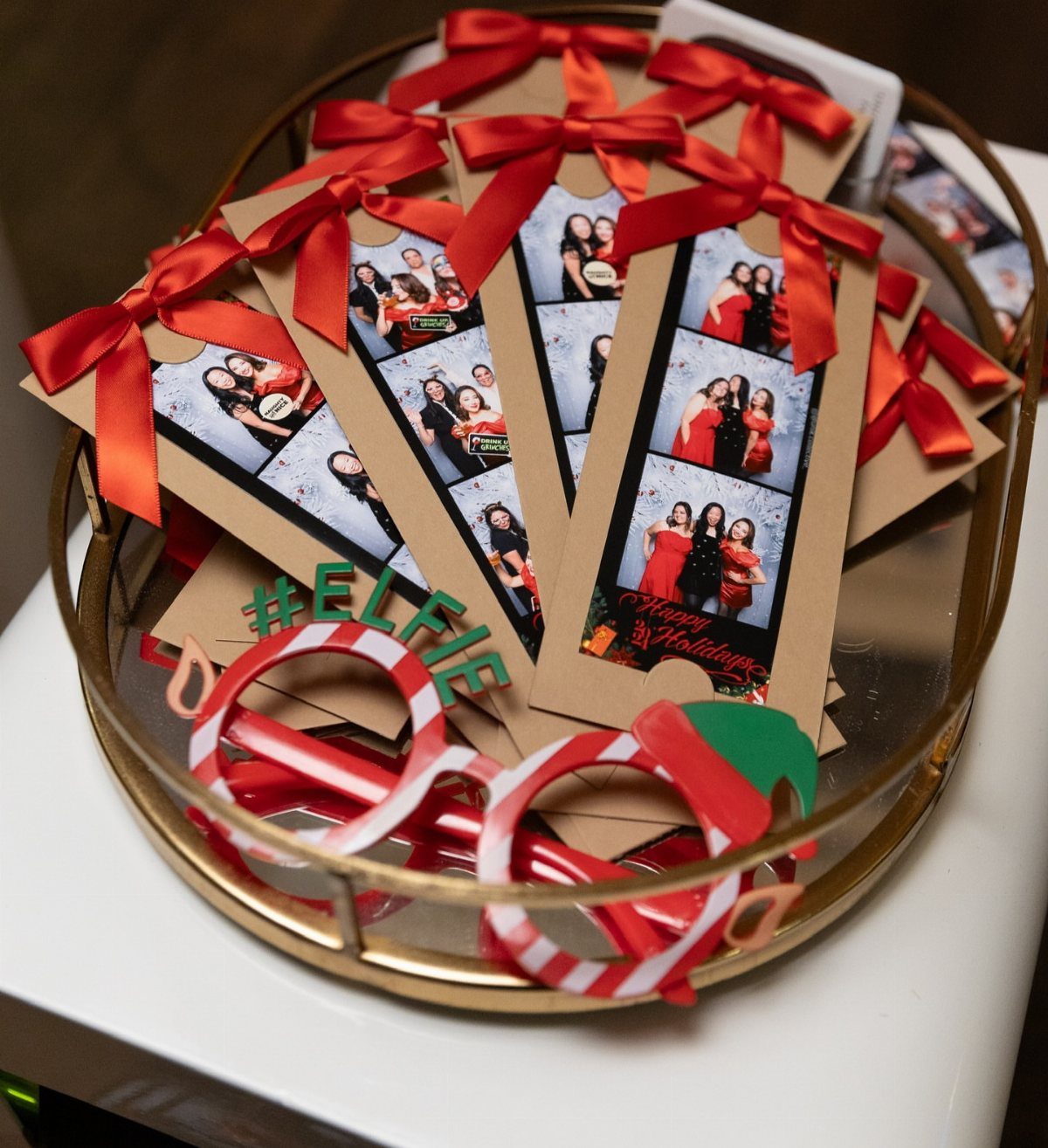 Photo booth strips with red ribbons and candy cane glasses on a gold tray.