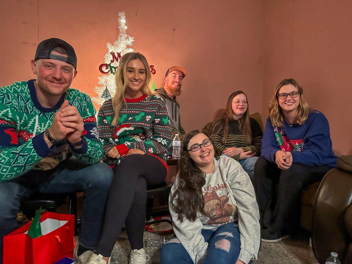 Five adults wearing festive holiday sweaters posing in a room with a white tree.