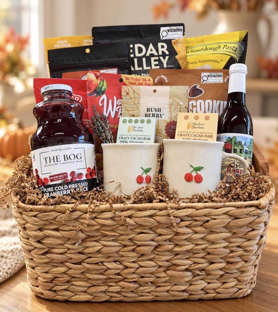 Woven gift basket filled with gourmet snacks, cranberry juice, and cherry-themed mugs.
