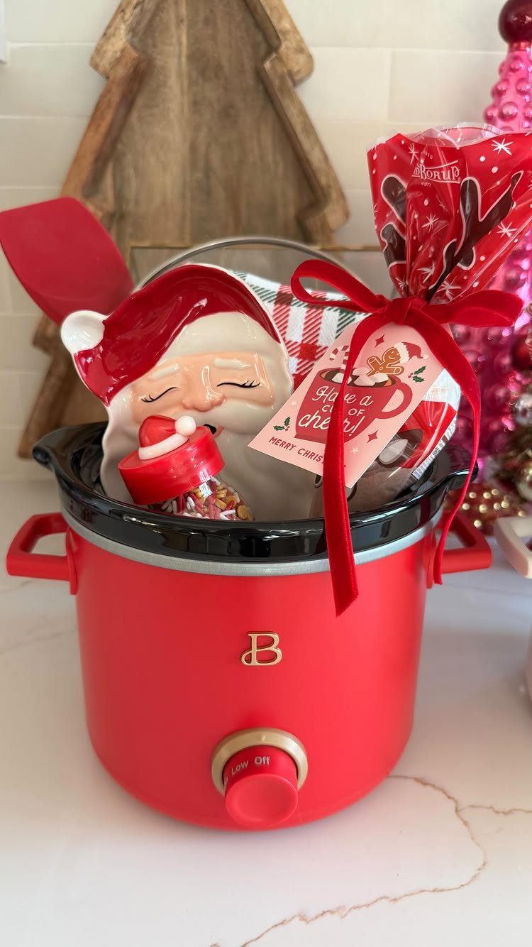 Red slow cooker filled with Christmas gifts, Santa figurine, and a red ribbon.