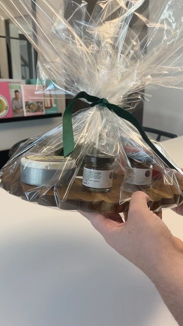 Wooden platter gift basket with cheese and jars wrapped in clear cellophane.