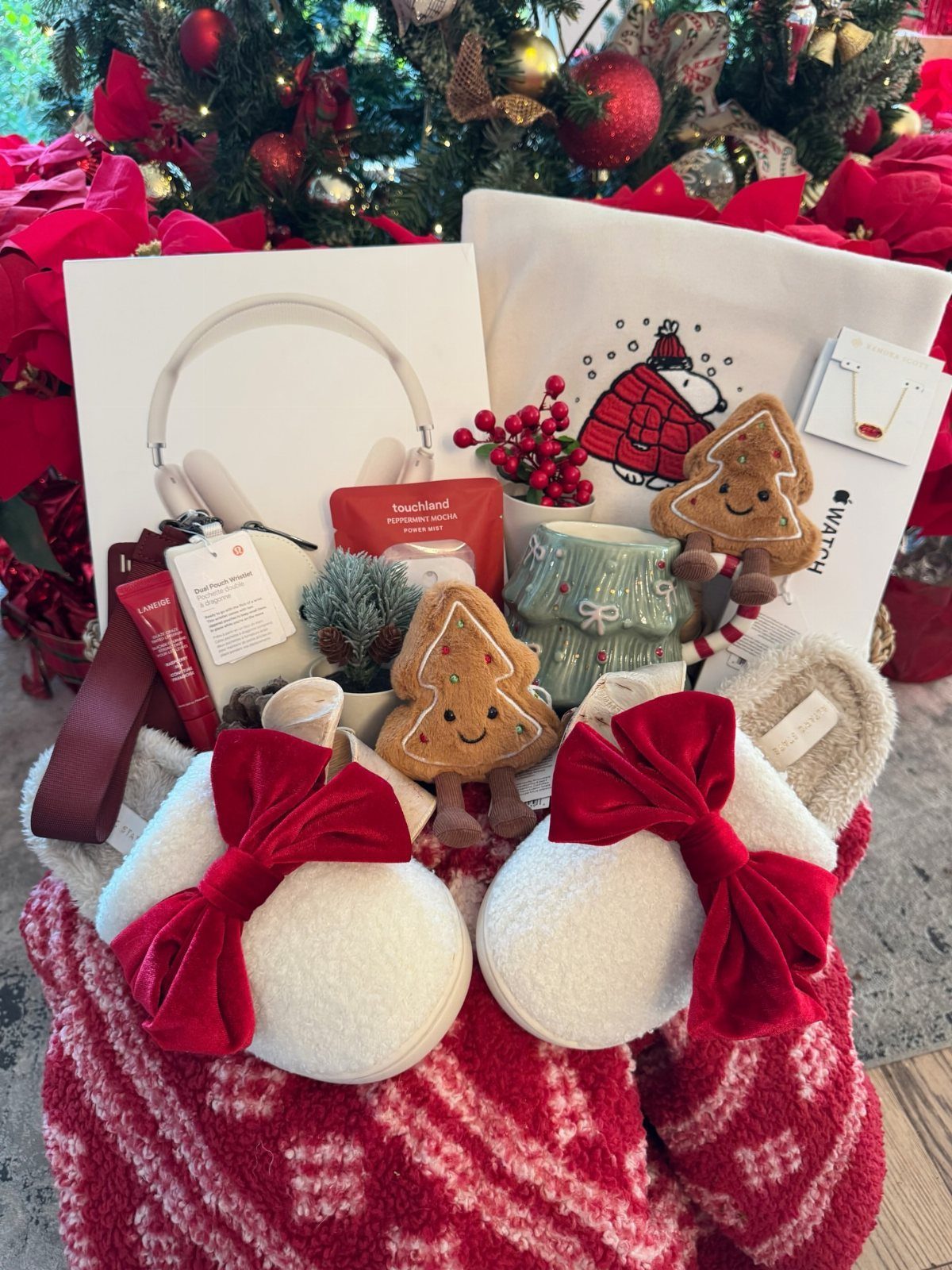 Festive gift hamper with white slippers, headphones, and gingerbread plushies by a Christmas tree.