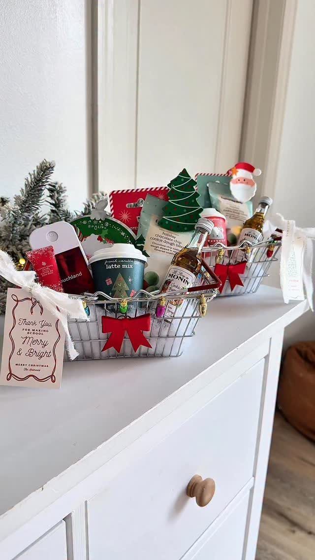 Two wire gift baskets with wine, coffee, and holiday decorations on a white dresser.