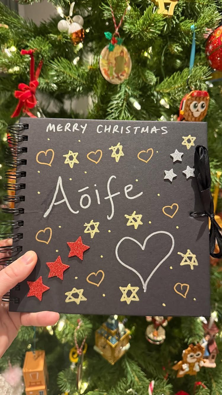 Personalized black Christmas notebook with glittery stars and hearts held before a tree.
