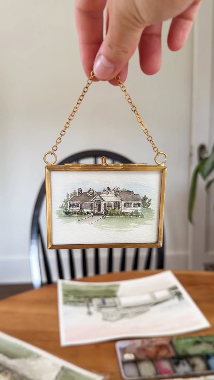 Gold-framed watercolor painting of a house held as a personalized gift pendant