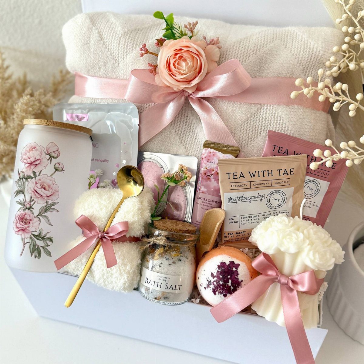 Spa gift set with white towel, floral candle, bath salts, and tea packets.
