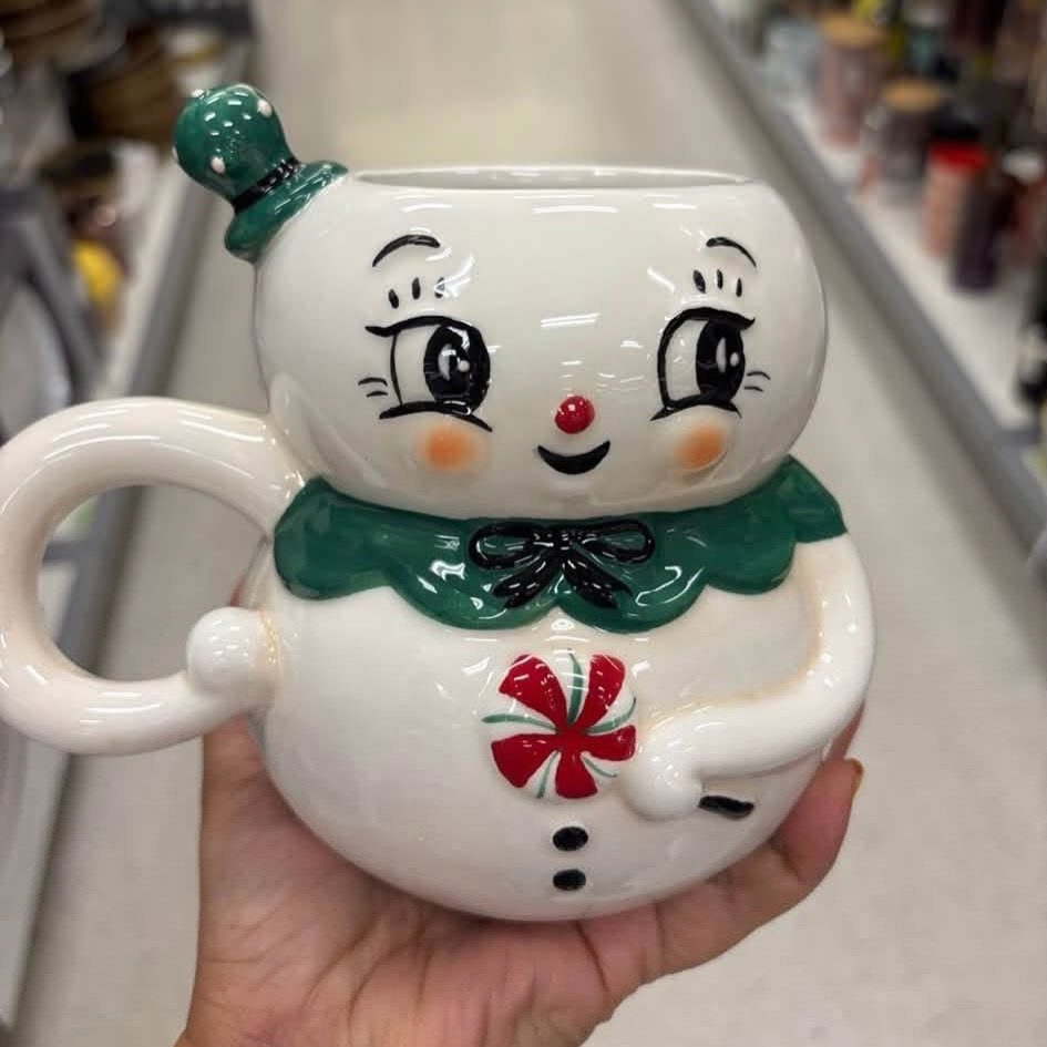 Snowman-shaped ceramic mug with a green collar and red peppermint button held in hand.