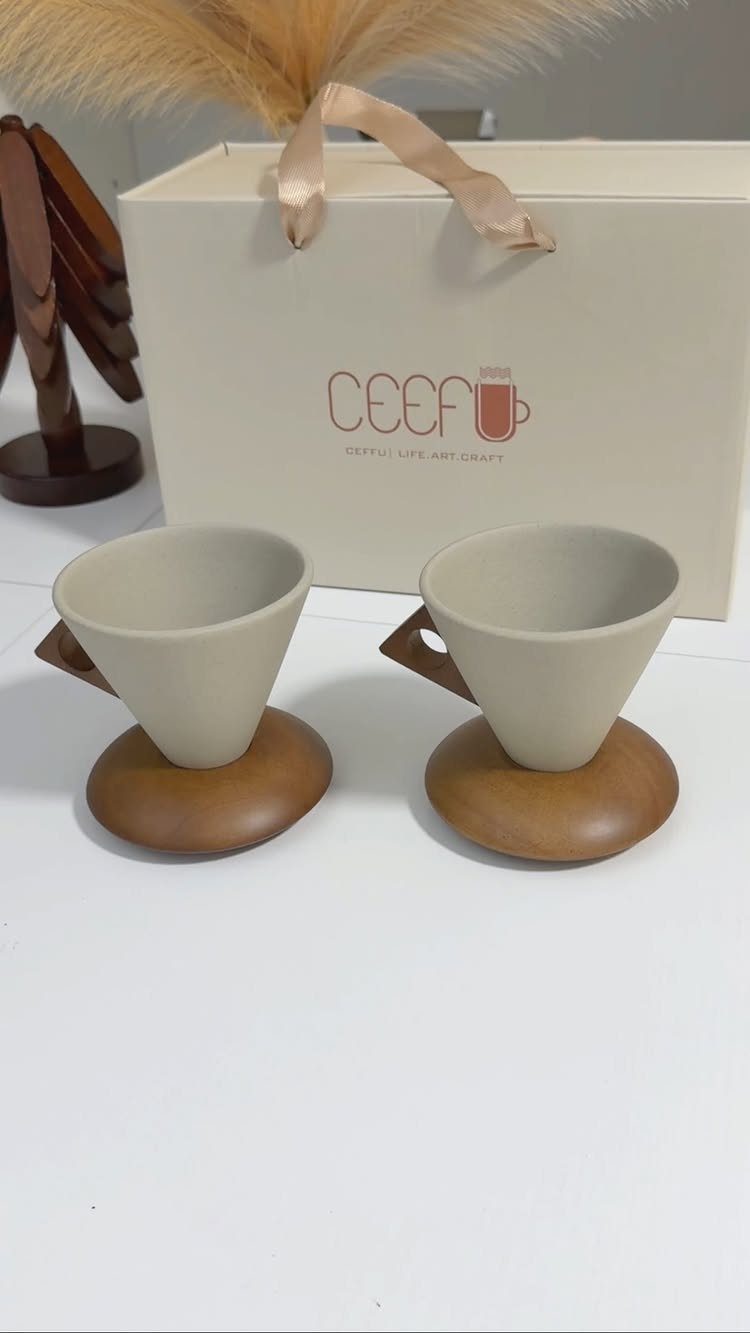 Two ceramic coffee drippers with wooden bases and handles next to a gift box.