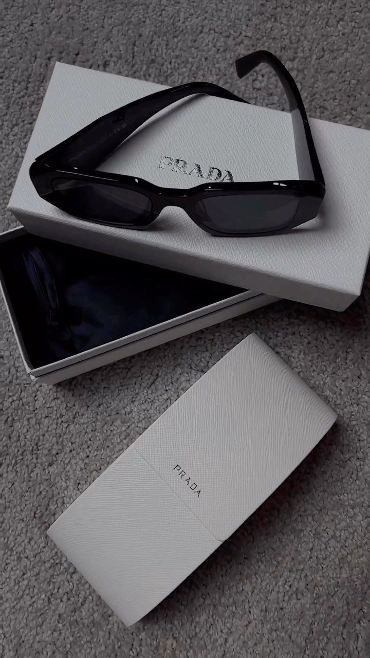 Black Prada sunglasses on a white branded box and case over grey carpet.