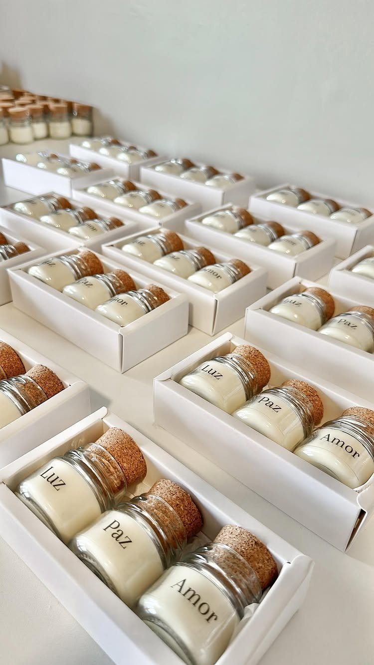 Small white candles in glass jars with cork lids arranged in white boxes.