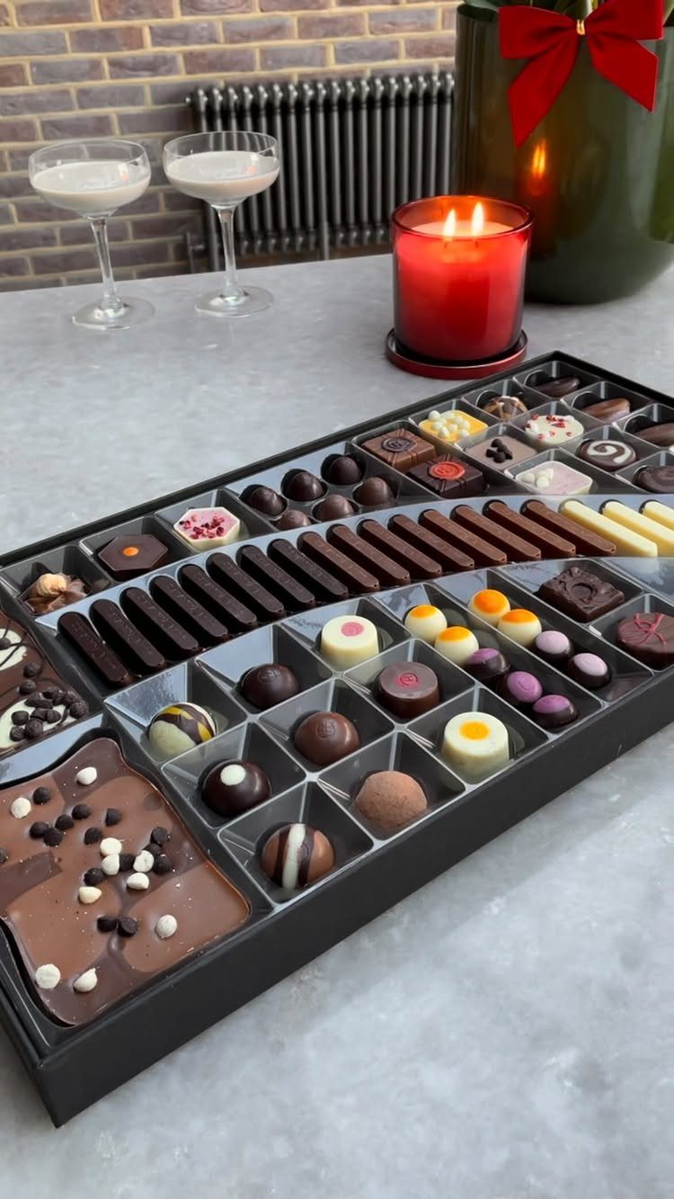Assorted chocolates in a black gift box with a red candle and drinks.