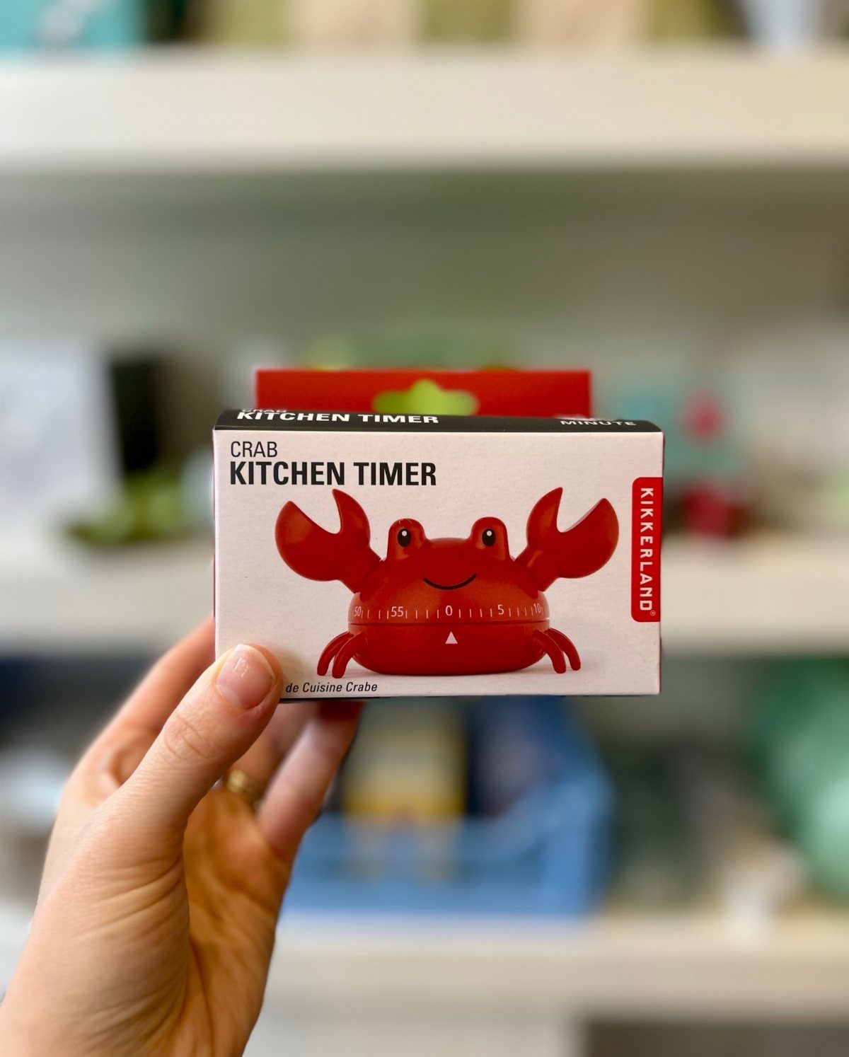 Hand holding a boxed red crab kitchen timer against a blurred shelf background