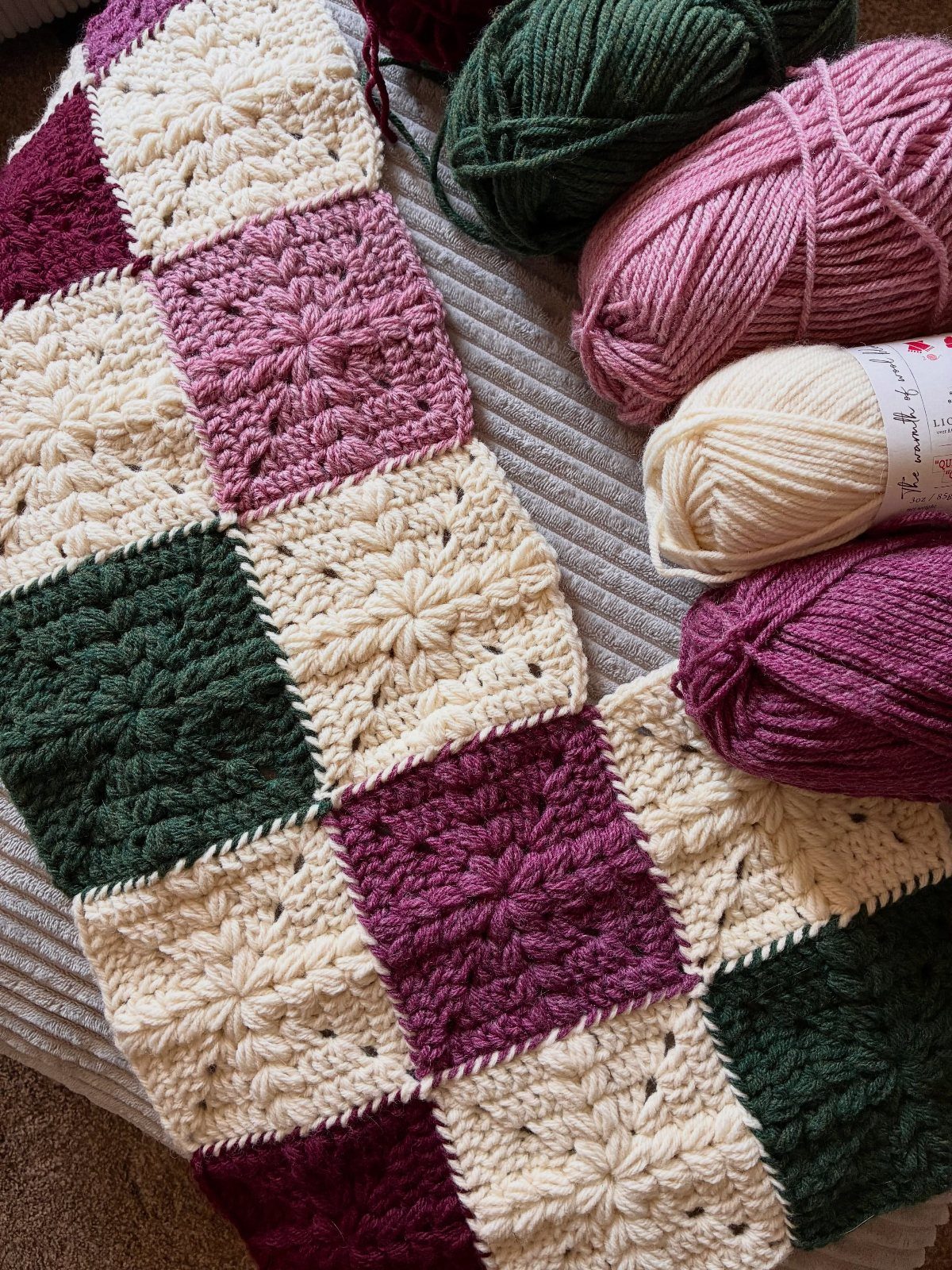 Crocheted granny square blanket in cream, green, and burgundy with matching yarn balls.