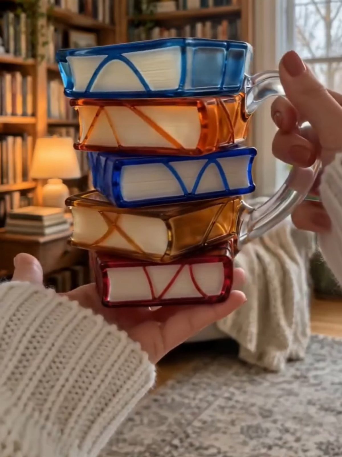 Stack of colorful book-shaped glass mugs held by hands in a cozy room