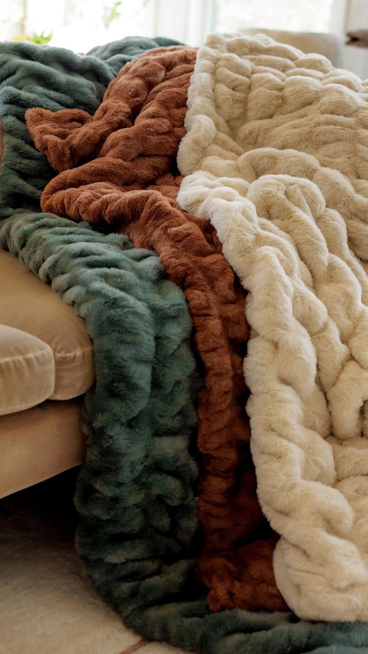 Three chunky knit blankets in cream, brown, and teal draped over a sofa.