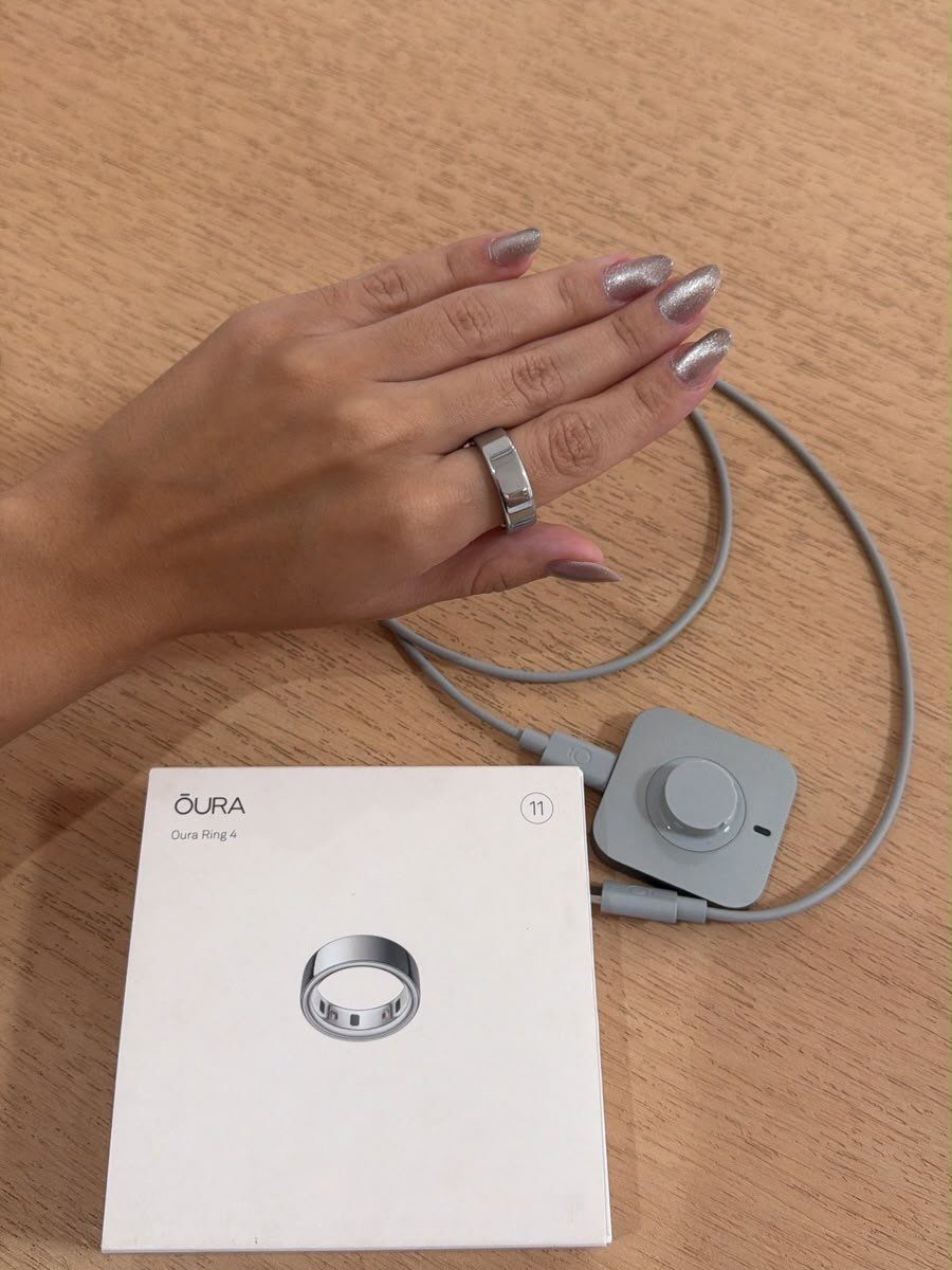 Hand wearing a silver smart ring next to its white packaging and charger.