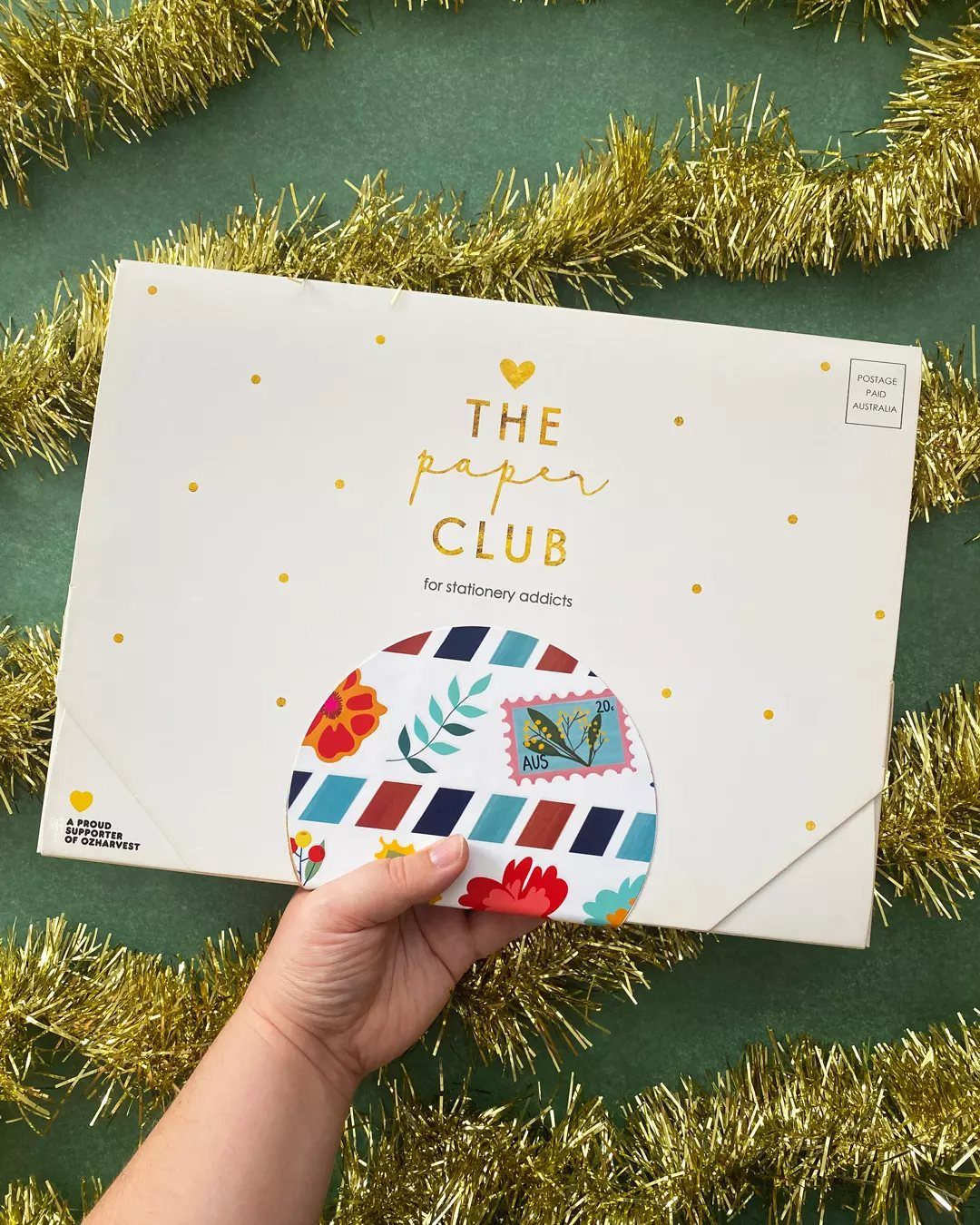 White subscription box envelope from The Paper Club held against gold tinsel background.