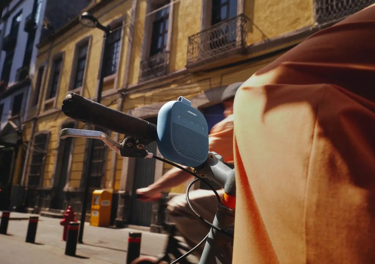 Blue portable speaker mounted on bicycle handlebars with a person in orange jacket.