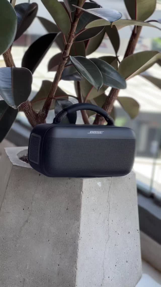Black Bose portable Bluetooth speaker sitting on a concrete pedestal with green leaves behind.