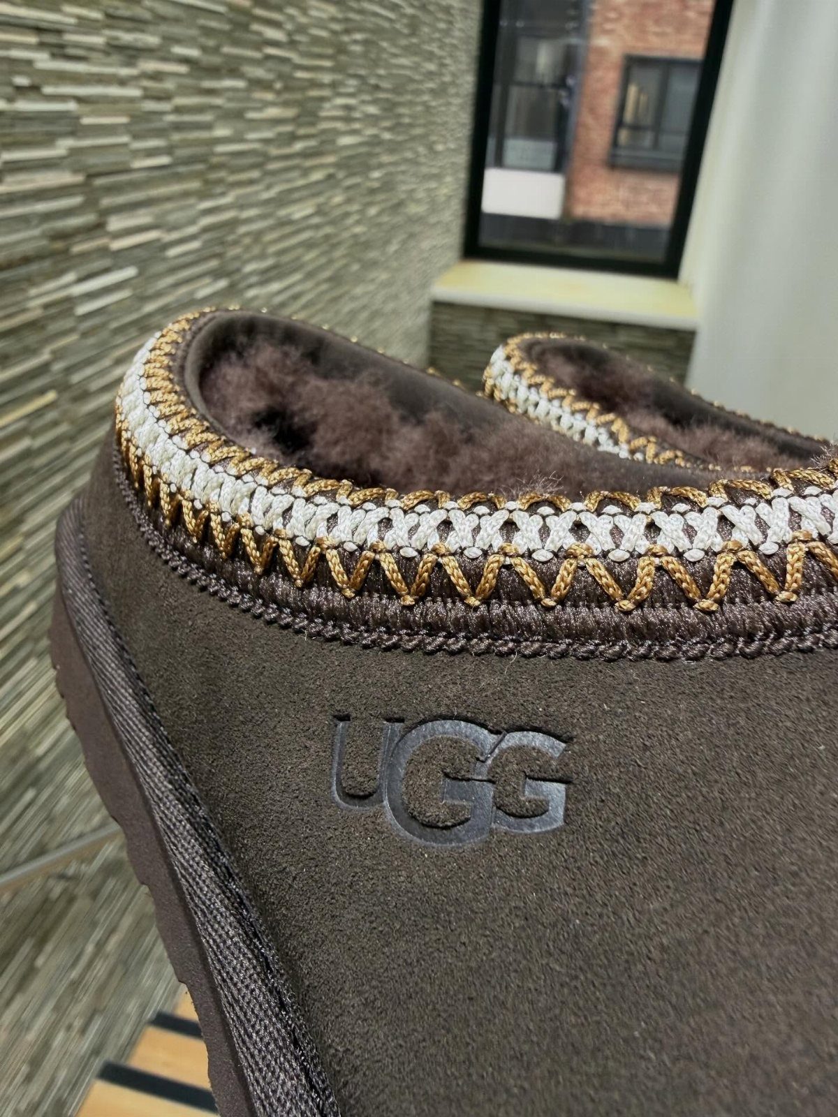 Brown suede UGG slippers with braided trim and faux-fur lining on stone background