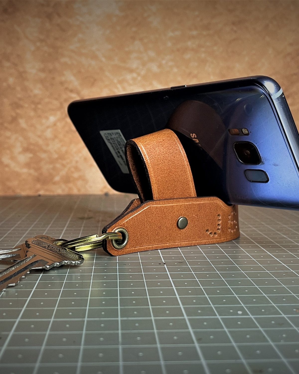 Blue smartphone on a brown leather stand with keys on a cutting mat