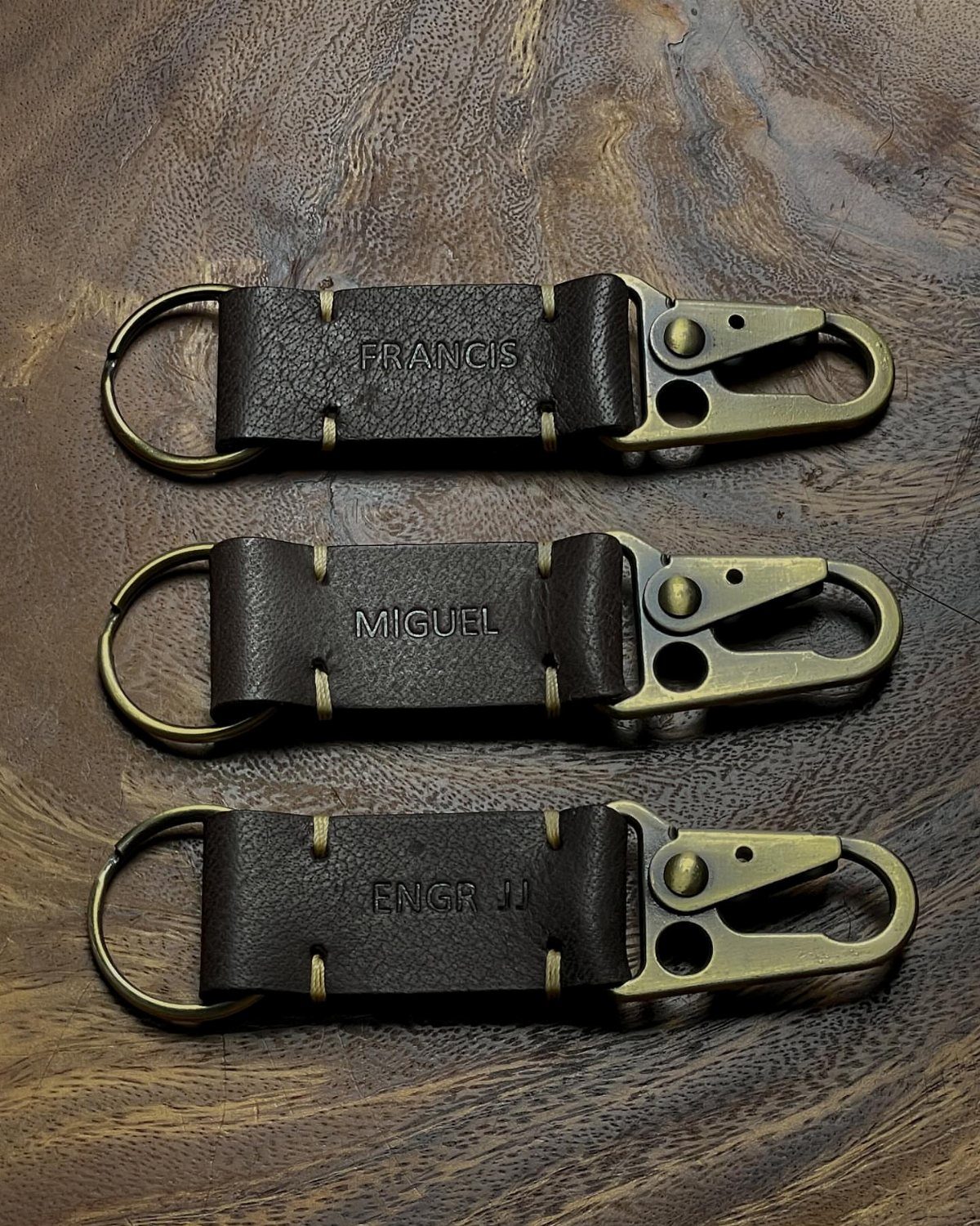 Three personalized leather key holders with brass clips on a dark wooden background.