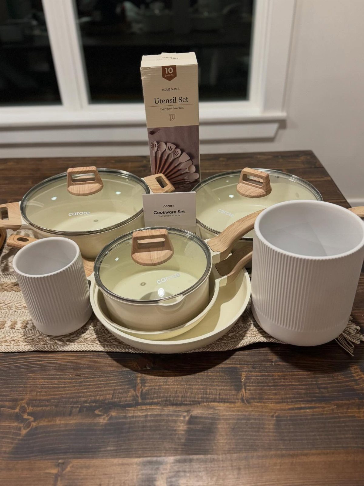 Cream-colored cookware set with pots, pans, and white containers on a wooden table.