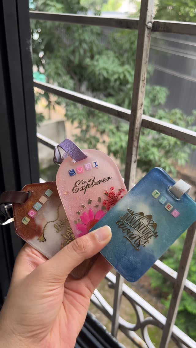 Three personalized luggage tags in pink, blue, and brown held by a hand.