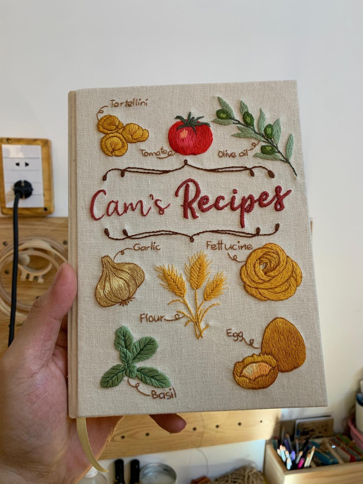 Embroidered linen recipe book featuring food illustrations and the text Cam's Recipes.