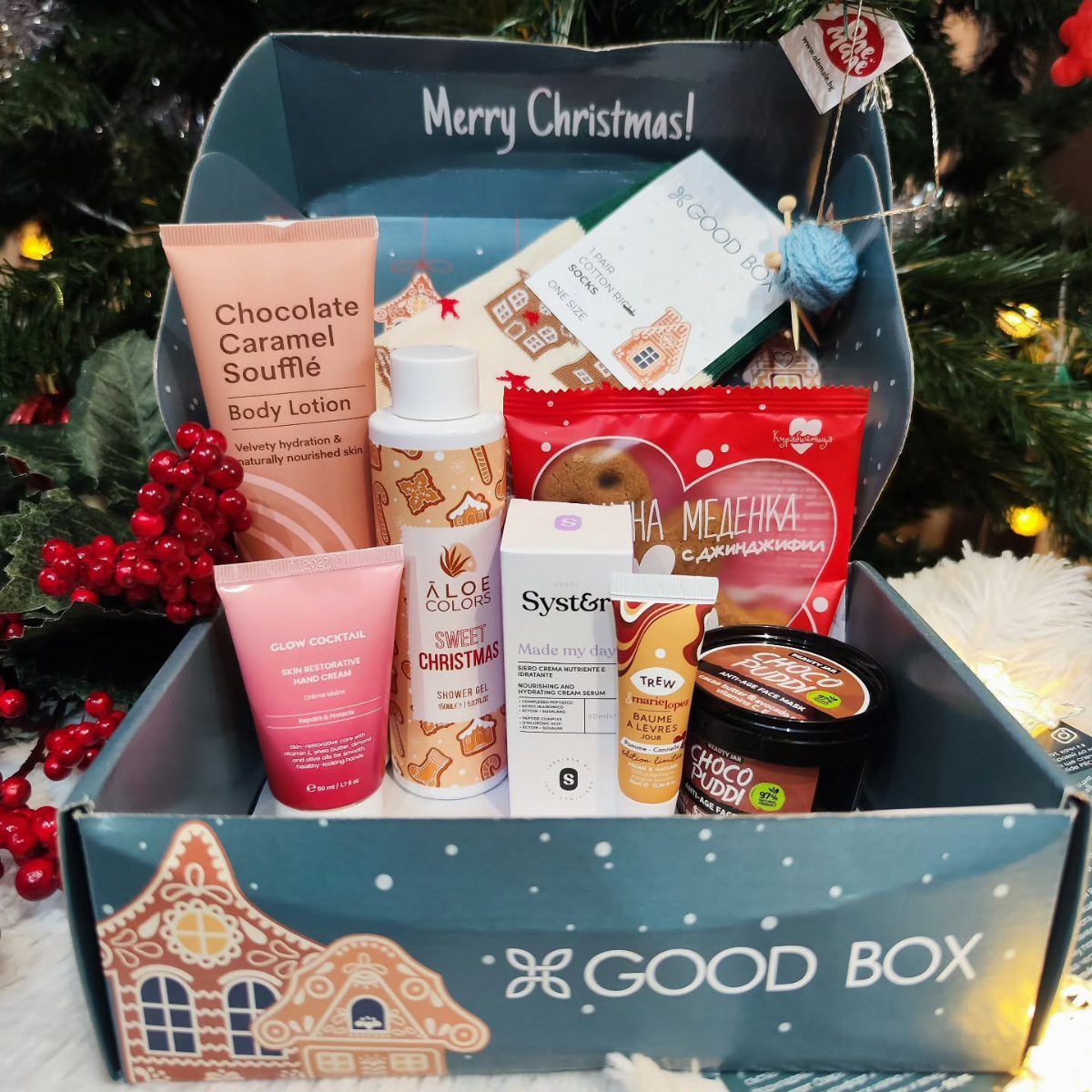 Blue gift box filled with skincare products and treats near a Christmas tree.