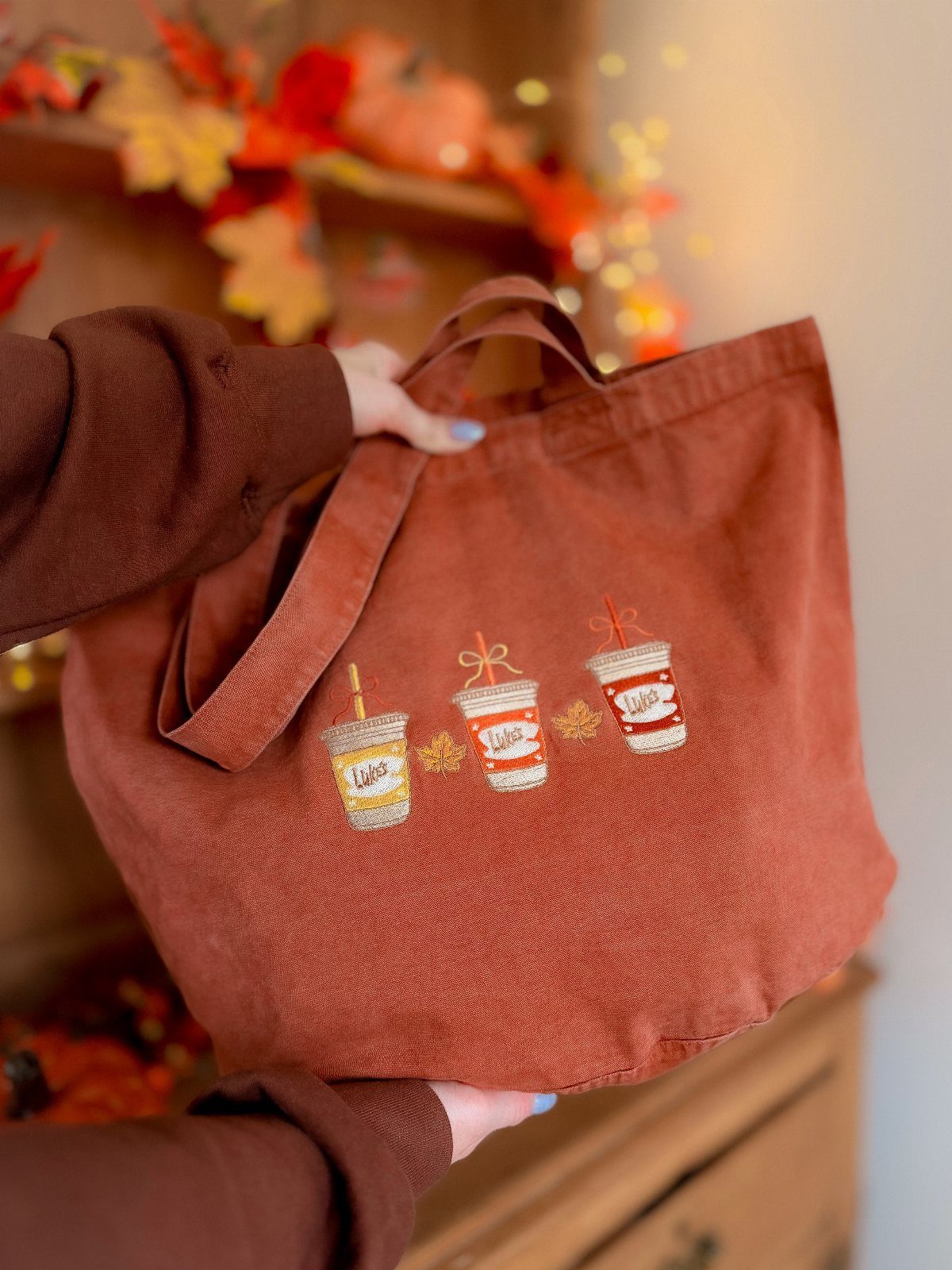 Rust-colored fabric tote bag with embroidered coffee cups held by a person.