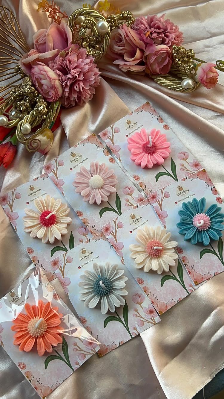 Flower-shaped candles in various colors arranged on floral cards over satin fabric.
